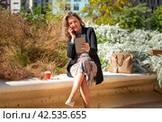 Smiling business woman with coffee sitting talking on a smartphone in hand outdoors. Стоковое фото, фотограф Raquel Corcoles Mendez / Фотобанк Лори