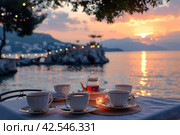 Table with teacups in front of a stunning sunset above the tranquil sea with mountains. Стоковое AI-изображение, AI-художник Юрий Поздников / Фотобанк Лори