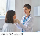 Middle-aged woman doctor wearing gloves checking patients sore throat or thyroid glands. Стоковое фото, фотограф Natalia Gdovskaia / Фотобанк Лори