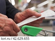 The hands of an unrecognizable older man hold a sales receipt next to a grocery cart in a supermarket. Стоковое фото, фотограф Владимир Федоров / Фотобанк Лори