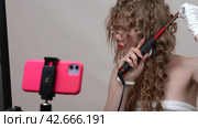 A woman blogger is using a curling wand to style hair, manually wrapping strands of blonde hair around the wand, recording herself to make a video blog using phone (2024 год). Редакционное видео, видеограф А. А. Пирагис / Фотобанк Лори