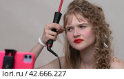 The blogger is using a curling wand to style hair by manually wrapping long hair around the wand. Woman sharing the process with us through a live stream on phone (2024 год). Редакционное видео, видеограф А. А. Пирагис / Фотобанк Лори