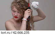 A blond young woman is using a curling tongs to style her long hair, manually wrapping sections of blonde hair around the iron wand to create thin curls (2024 год). Редакционное видео, видеограф А. А. Пирагис / Фотобанк Лори