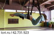 A woman of European appearance is engaged in aerial yoga, performs the exercise Supta Baddha Konasana, the pose of a resting goddess, training in sportswear in the studio. Стоковое фото, фотограф Сергей Чайко / Фотобанк Лори