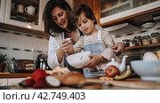 A young mother shows her little daughter how to make pie dough in the kitchen. The continuity of traditions in generations. Стоковое AI-изображение, AI-художник Ярославский Максим / Фотобанк Лори