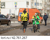 В Москве на улице встретились два курьера сервисов еды. Курьер сервиса "Яндекс.Еда" идет пешком к клиенту, а велокурьер  в форменной одежде "Сбермаркет". сидя на своем велосипеде, отдыхает со смартфоном в руках (2024 год). Редакционное фото, фотограф Наталья Николаева / Фотобанк Лори