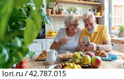 An elderly man shows his wife vacation photos on his smartphone. They have breakfast in the kitchen in the morning. Стоковое AI-изображение, AI-художник Ярославский Максим / Фотобанк Лори