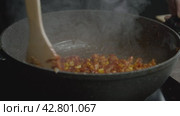 The cook combines chickpeas, corn, and chicken with a variety of spices in a large, black wok using a wooden spoon. Стоковое видео, видеограф Strunov David / Фотобанк Лори