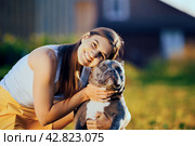 Dog is forced to look at camera with its head forcibly lifted, smiling portrait of young European woman with an American Bully in countryside at end of summer day. Стоковое фото, фотограф Владимир Григорьев / Фотобанк Лори