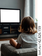 Rear view of little girl kid 5-6 year old watching tv sits on armchair looking cartoon. Adorable dependency girl child, empty isolated screen. Television addiction tv concept. Copy ad text space. Стоковое AI-изображение, AI-художник Aleksandr Golubev / Фотобанк Лори