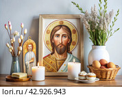 Orthodox Easter. Easter cake, painted eggs, icons, faces of saints, flowers, church candles and Easter decor on a wooden table in an old village house. Стоковое AI-изображение, AI-художник Photojuli / Фотобанк Лори