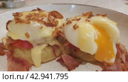Close up of Eggs Benedict with liquid egg yolk running out. Two poached eggs on potato pancakes or toasted bread with tomatoes, fried onions and bacon with hollandaise sauce. Стоковое видео, видеограф Светлана Ельцова / Фотобанк Лори