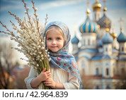 Palm Sunday. Christianity. Portrait of a three-year-old girl in a Russian folk shawl with willow branches in her hands against the background of a church and a sunset. Стоковое AI-изображение, AI-художник Photojuli / Фотобанк Лори