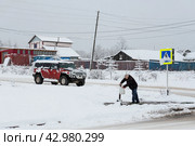 Мужчина набирает воду из водоразборной колонке на окраине северного города. По заснеженной проезжей части едет большой внедорожный автомобиль. Проблемы централизованного водоснабжения в регионах России. Холодная снежная погода. (2023 год). Редакционное фото, фотограф Андрей Степанов / Фотобанк Лори