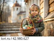 Happy bright Easter. Christianity. Portrait of a three-year-old girl in a Russian folk dress and shawl with a wicker basket with Easter eggs in her hands against the background of an Orthodox church. Стоковое AI-изображение, AI-художник Photojuli / Фотобанк Лори