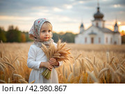 Russian Russian girl in a white dress and a Russian headscarf holds golden ears of wheat against the background of a ripening field and an Orthodox church. The concept of planting and harvesting a rich harvest. Rural landscape. Стоковое AI-изображение, AI-художник Photojuli / Фотобанк Лори