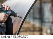 A man holds a battery-powered screwdriver and assembles a new greenhouse made of polyethylene, polycarbonate. Macro photography. Side view. Preparing for the spring garden season. Стоковое фото, фотограф Photojuli / Фотобанк Лори