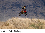 Motocross rider in orange and black gear performing a mid-air jump over a hill in Wanaka, New Zealand, against a mountainous backdrop. (2024 год). Редакционное фото, фотограф Евгений Дробитько / Фотобанк Лори