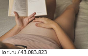 Беременная женщина в расслабленном состоянии лежит и увлеченно читает книгу. Она поглаживает рукой свой живот. На ней ребристый бежевый топ. Акцент на ее животе и книге. Стоковое видео, видеограф Воловодов Владимир / Фотобанк Лори