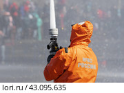 Fireman in an orange protective uniform pours powerful stream of water upward from fire hose. Inscription on back of firefighter is in Russian EMERCOM of Russia (2019 год). Редакционное фото, фотограф А. А. Пирагис / Фотобанк Лори