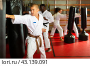Portrait of concentrated man in kimono practicing powerful body punches on heavy punchbag during workout. Стоковое фото, фотограф Яков Филимонов / Фотобанк Лори