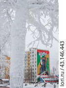 Город Тында, Амурская область, Россия. Вид на большое граффити на стене жилого дома, посвящённое строителям Байкало-Амурской магистрали (БАМ) и города Тынды. Мурал с изображением молодых энергичных людей, идущих на стройку. На заснеженной улице скульптурная композиция в виде серпа и молота. На переднем плане, покрытый инеем, ствол дерева. (2024 год). Редакционное фото, фотограф Андрей Степанов / Фотобанк Лори