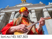 Мужчина в традиционном русском народном костюме играет на балалайке на фоне арки Центрального входа на выставочный комплекс ВДНХ в городе Москве, Россия (2024 год). Редакционное фото, фотограф Николай Винокуров / Фотобанк Лори
