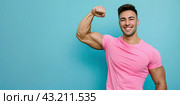 A young man in a pink T-shirt shows his biceps on his right arm on a light blue background. Copy spase. Стоковое AI-изображение, AI-художник Ярославский Максим / Фотобанк Лори