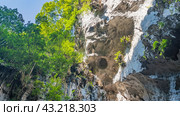 A fragment of a sheer limestone cliff. Caverns, furrows, and crevices are visible. There is green vegetation on the slopes. Bottom view. Tree branches against a clear blue sky. Malaysia. Batu caves. Стоковое фото, фотограф Вера Ивановна Кубарева / Фотобанк Лори