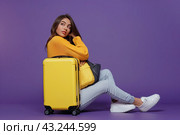 A thoughtful traveler girl leans against a yellow suitcase on a uniform lilac background. Copy space. The concept of waiting for a vacation. Стоковое AI-изображение, AI-художник Ярославский Максим / Фотобанк Лори
