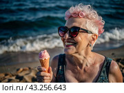 Portrait of an extraordinary, stylish, fashionable elderly woman, a pensioner of 70 years old, with ice cream in her hands on vacation on the beach by the sea. The concept of happy seniors and delicious desserts. Стоковое AI-изображение, AI-художник Photojuli / Фотобанк Лори
