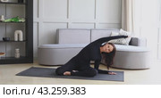 A middle-aged woman performs side bends while sitting in a half-lotus position, stretching the core muscles, training in sports overalls in the room, full-length view. Стоковое видео, видеограф Сергей Чайко / Фотобанк Лори
