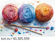 Three balls of yarn of different colors: pink, blue and orange, located on a white background. There are bright spots of paint around the balls, creating a feeling of artistic chaos and creativity. Next to the yarn are two wooden knitting needles with small balls at the ends, emphasizing the theme of knitting and needlework. The painting is made in a watercolor style with an emphasis on bright and rich colors, creating a warm and cozy atmosphere. Стоковое AI-изображение, AI-художник Анна Кривицкая / Фотобанк Лори