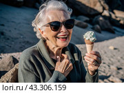 Portrait of an extraordinary, stylish, fashionable elderly woman, a pensioner of 70 years old, with ice cream in her hands on vacation on the beach by the sea. The concept of happy seniors and delicious desserts. Стоковое AI-изображение, AI-художник Photojuli / Фотобанк Лори
