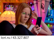 Милая девушка-подросток в очках сидит и смотрит на дисплей смартфона на фоне размытых гирлянд. Стоковое фото, фотограф Валуйских Владимир / Фотобанк Лори