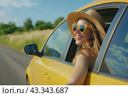 A happy girl in glasses and a straw hat leans out of the window of a yellow car towards the sun. Copy space. Стоковое AI-изображение, AI-художник Ярославский Максим / Фотобанк Лори