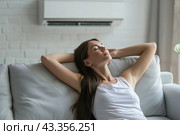 Young woman suffering during a heat heat sitting under air conditioner at home resting on a sofa. Exhausted girl feeling sweaty switching on modern AC system. Hot summer. Fresh clean air in apartment. Стоковое AI-изображение, AI-художник Марина Демидюк / Фотобанк Лори