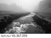 Monochrome landscape of a mountain river flowing through a narrow gorge. The banks of the river are covered with stones, and in the background you can see majestic cliffs lost in thick fog. The river flows rapidly, the water foams and creates a feeling of movement. The scene conveys an atmosphere of mystery and the harsh beauty of nature, enhancing the drama of the landscape. Стоковое AI-изображение, AI-художник Анна Кривицкая / Фотобанк Лори