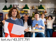 Company of upset young adult sports fans holding flag of England and worrying about defeat during game match on TV in pub. Стоковое фото, фотограф Яков Филимонов / Фотобанк Лори