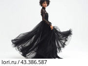 Young woman in an elegant black evening dress. The dress has a long, flowing hem made of translucent material, which creates the effect of lightness and airiness. The top of the dress is made of lace fabric with long sleeves, which gives the outfit sophistication and sophistication. The woman poses in motion, holding the hem of the dress, which emphasizes the beauty and dynamics of the outfit. She has dark curly hair and expressive makeup. The background of the image is neutral and light, which allows you to focus on the model and her outfit. Стоковое AI-изображение, AI-художник Анна Кривицкая / Фотобанк Лори