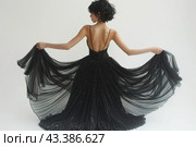 Young woman in an elegant black dress. She stands with her back to the camera, holding the edges of the dress in her hands, creating the effect of a fluttering train. The dress has a plunging back and is adorned with thin straps and sparkling rhinestones that add sophistication and sparkle. The background is clean and white, which emphasizes the contrast between the dress and the surrounding space, focusing attention on the figure and dress. Стоковое AI-изображение, AI-художник Анна Кривицкая / Фотобанк Лори