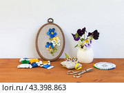 Handmade embroidery with satin ribbons of summer pansies in hoop, bouquet of fresh pansy flowers in vase on wooden table. Nearby is bobbins of satin ribbons, needles and scissors for work. DIY concept. Стоковое фото, фотограф Виктория Катьянова / Фотобанк Лори
