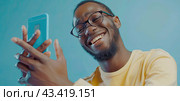A black young man looks at the smartphone screen with a smile. Blue background. Стоковое AI-изображение, AI-художник Ярославский Максим / Фотобанк Лори