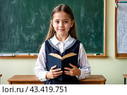 First grader girl in school uniform reading book at blackboard in classroom, interest look. Adorable child schoolgirl with book posing inside. School learning, education concept. Copy ad text space. Стоковое AI-изображение, AI-художник Aleksandr Golubev / Фотобанк Лори