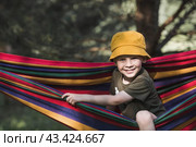 Summer holidays for children in the fresh air.. A cute little boy is having fun with a colorful hammock in the backyard. A child in a hammock. Стоковое фото, фотограф Photojuli / Фотобанк Лори