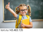 A cute girl with glasses shows the class with her hand. The child goes to school for the first time. A child with a school bag and a book. A child in the classroom on the background of a blackboard. Back to school. Стоковое AI-изображение, AI-художник Photojuli / Фотобанк Лори
