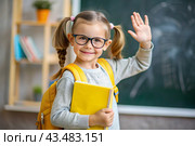 A cute girl with glasses shows the class with her hand. The child goes to school for the first time. A child with a school bag and a book. A child in the classroom on the background of a blackboard. Back to school. Стоковое AI-изображение, AI-художник Photojuli / Фотобанк Лори