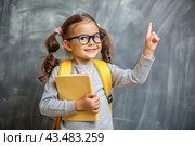 A cute girl with glasses shows the class with her hand. The child goes to school for the first time. A child with a school bag and a book. A child in the classroom on the background of a blackboard. Back to school. Стоковое AI-изображение, AI-художник Photojuli / Фотобанк Лори