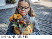 A pretty little girl with glasses and a bouquet of autumn flowers goes to school for the first time. A child with a school bag, a book and flowers. A child on a city street. Back to school. Стоковое AI-изображение, AI-художник Photojuli / Фотобанк Лори
