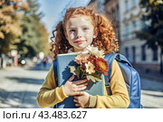 A pretty red-haired girl with glasses and a bouquet of autumn flowers goes to school for the first time. A child with a school bag, a book and flowers. A child on a city street. Back to school. Стоковое AI-изображение, AI-художник Photojuli / Фотобанк Лори